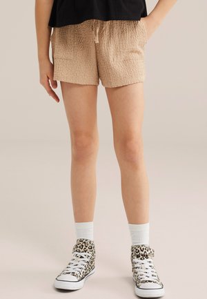 Nogi w beżowych teksturowanych szortach i białych skarpetkach, w panterkowych sneakersach na wysokiej podeszwie, na jednolitym tle.