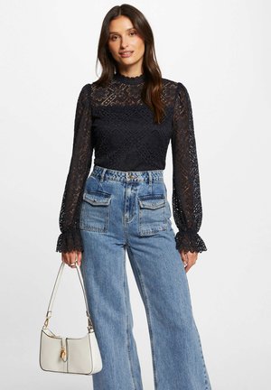 Top lungo in pizzo nero con maniche lunghe e scollo alto, motivo floreale dettagliato e orli arricciati, abbinato a jeans blu a vita alta e larghi, e una borsa bianca.