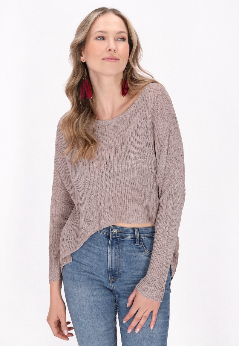 Pulover din tricot ribetat, de culoare maro deschis, cu mâneci lungi, față scurtă și slituri laterale, purtat cu blugi albaștri. Cerceii din pene roșii adaugă detaliu.