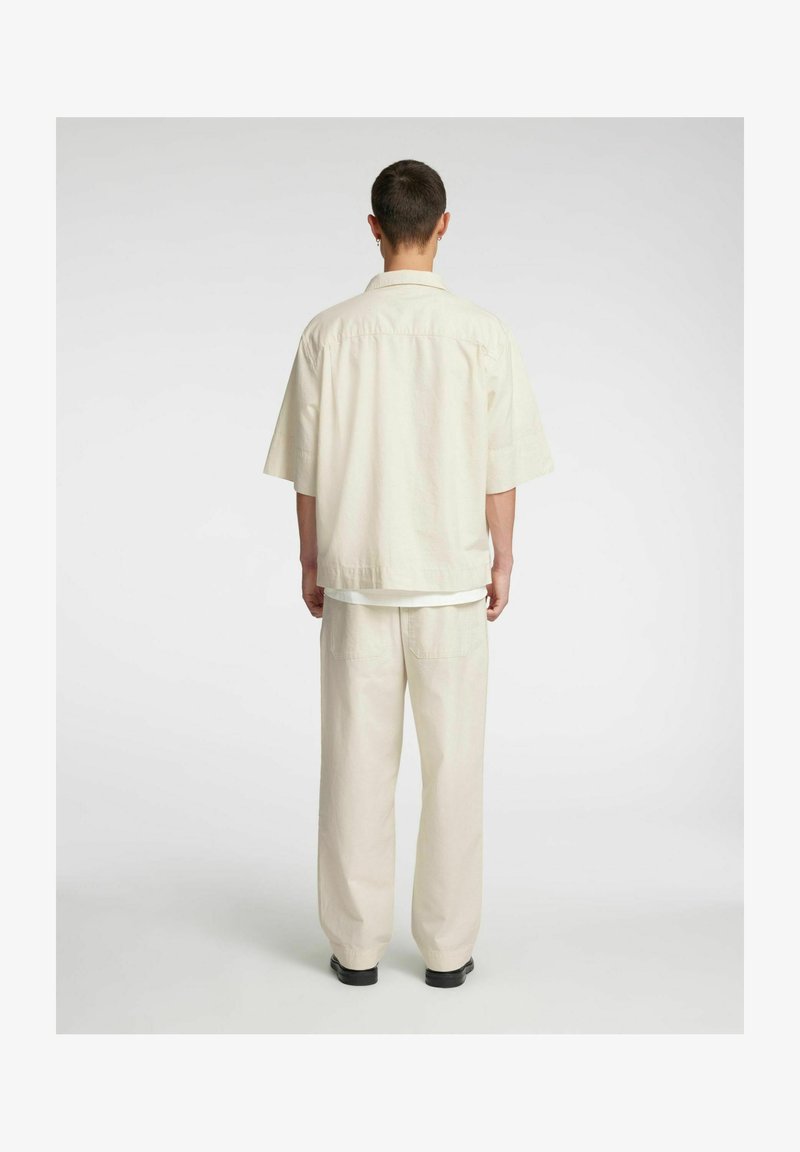 Lys beige oversized skjorte med slagkrage, korte ermer og matchende lyse bukser. Antrekket er laget av mykt, teksturert stoff.