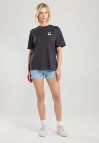 Harmaa puuvillainen t-paita, jossa on lyhyet hihat ja neliön muotoinen vihreä ja keltainen logo rinnassa. Yhdistetty vaaleansinisiin farkkushortseihin ja valkoisiin tennareihin.