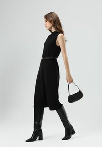 Zwarte mouwloze jurk met een hoge halslijn en een ceintuur in de taille, gecombineerd met zwarte kniehoge laarzen en een klein zwart handtasje.