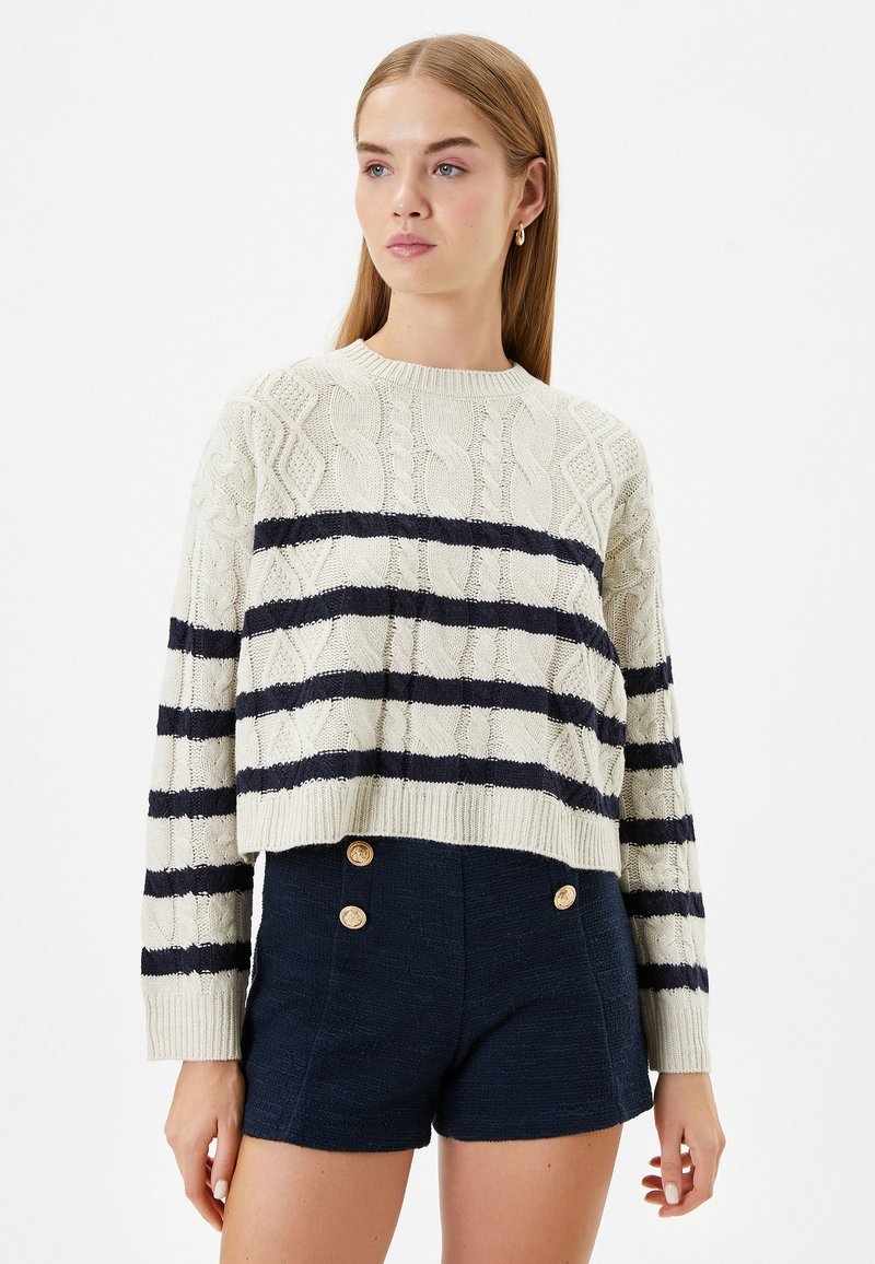 Crème kabelgebreide trui met marineblauwe horizontale strepen, ronde halslijn en lange mouwen. Draag hem met donker marineblauwe hoge taille shorts met gouden knopen.