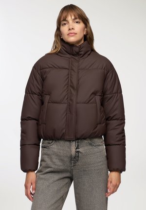 Giacca puffer marrone con colletto alto, motivo trapuntato e design accorciato, abbinata a jeans grigi chiari con una stampa sottile.