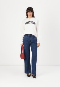Hvit langermet genser med svart "TOMMY JEANS" tekst, kombinert med blå vide jeans og svarte spisse sko, holder en rød veske.