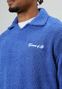 Blauwe, zachte fleece trui met een kraag, voorzien van een geborduurd logopatch in wit op de linkerborst. Gestructureerde, zachte stof.