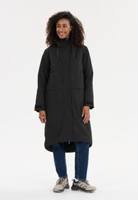 Manteau long noir avec capuche, doté d'une coupe droite et de deux poches avant. Porté sur un jean bleu et des baskets grises et noires.