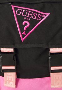 Svart ryggsäck med en rosa broderad triangel-logotyp med "GUESS" och ett frågetecken, justerbara rosa axelremmar och plastspännen.