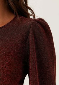Röd glittrig klänning med figurnära liv och puffärmar, tillverkad av textil med struktur som fångar ljuset och skapar en skimrande effekt.