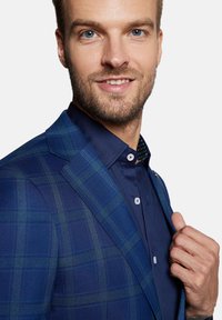 Niebieska marynarka w kratę z teksturowanej tkaniny i wyraźnie zarysowanymi ramionami, w zestawieniu z ciemnoniebieską koszulą z wzorzystym kołnierzykiem.