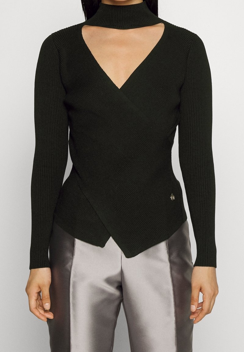 Femme portant un haut noir à manches longues côtelé avec un col haut et une découpe en V profonde, assorti à un pantalon gris brillant.