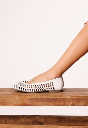 Gamba con scarpa piatta bianca con motivo a ritagli rettangolari e cinturino beige, appoggiata su una superficie di legno con sfondo chiaro.