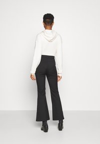 Valkoinen huppari ja mustat leveälahkeiset housut, joissa on sileä pinta. Housuissa on korkeavyötärö ja mallilla on jalassaan tukevat mustat saappaat.