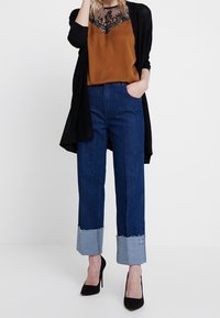 Brun sidentopp med svart spetsdetalj, svart kofta, högmidjade blå denimjeans med fransade ljusblå manschetter, svarta högklackade skor.