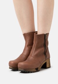 Stivali ankle in pelle marrone con cerniera laterale, punta arrotondata e un tacco in legno testurizzato. Dettagli cuciti lungo i bordi.