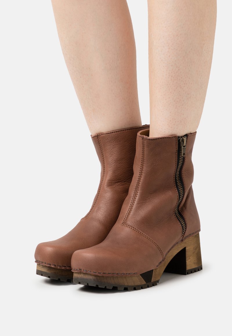 Stivali ankle in pelle marrone con cerniera laterale, punta arrotondata e un tacco in legno testurizzato. Dettagli cuciti lungo i bordi.
