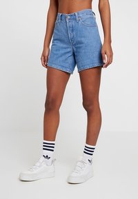Persoon die mid-lengte blauwe spijkershorts draagt, witte sneakers en witte kousen met marineblauwe strepen tegen een effen achtergrond.