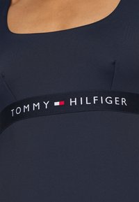 Costume da bagno blu navy con scollatura a forma di barca e tessuto morbido. Presenta un elastico nero con la scritta "TOMMY HILFIGER" in bianco, rosso e blu.
