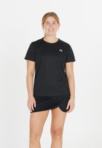 Nuori nainen hymyilee seisten, päällään musta lyhythihainen urheilupaita, jossa on "FZ"-logo, sekä mustat urheiluhameet valkoista taustaa vasten.