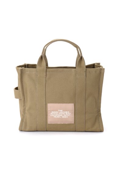 Saco de lona caqui com duas alças curtas, forma retangular e um rótulo rosa com texto branco. Tecido texturizado e costura em contraste.