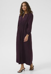 Lång lila klänning med smockad midja, V-ringning och långa ärmar. Tillverkad av lätt material, har en avslappnad silhuett och flödande design.