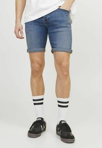 Pantalones cortos de mezclilla de color azul medio, con el dobladillo vuelto. Combinados con zapatillas negras y calcetines blancos con dos rayas negras.