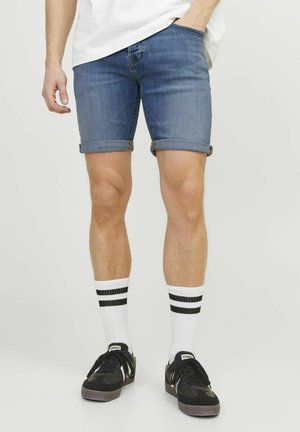 Pantalones cortos de mezclilla de color azul medio, con el dobladillo vuelto. Combinados con zapatillas negras y calcetines blancos con dos rayas negras.