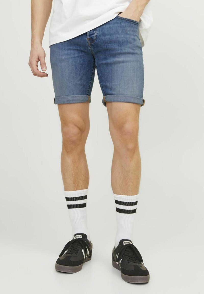 Pantalones cortos de mezclilla de color azul medio, con el dobladillo vuelto. Combinados con zapatillas negras y calcetines blancos con dos rayas negras.