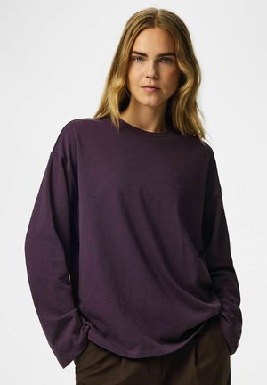 Femme aux longs cheveux blonds portant une chemise ample à manches longues violet foncé et un pantalon foncé, debout devant un fond clair uni.