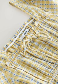 Žltá károvaná látka so vzormi modrých sŕdc a kvetov. Elastický pás s bielou etikou označenou "NEXT" a volánikovými okrajmi.