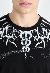Black cotton t-shirt featuring a white tribal-inspired graphic design with shapes resembling medical symbols and abstract patterns on the chest.