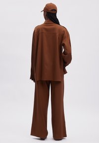 Chemise marron oversize à manches longues avec un empiècement au dos, portée avec un pantalon large assorti et une casquette marron. Tissu lisse et doux.
