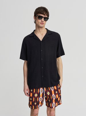 Uomo con camicia nera a maniche corte con bottoni, occhiali da sole neri e pantaloncini con motivo geometrico multicolore, in piedi con una mano in tasca.