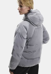 Szara kurtka puchowa z kapturem, o gładkiej fakturze, z elastycznymi mankietami i zamkniętą kieszenią po lewej stronie.