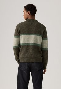 Strikkegenser i mørkegrønn med horisontale striper i lys grønn og krem. Har krage og teksturert overflate. Bæres med mørke jeans.