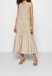 Robe sans manches beige avec une texture plissée. Ourlet évasé, longueur cheville. Fabriquée en tissu léger, elle possède un col rond.