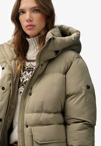 Chamarra tipo puffer en caqui con capucha. Presenta un diseño acolchado, botones a presión y un parche con el logo en la manga. Se lleva encima de un suéter de diseño.