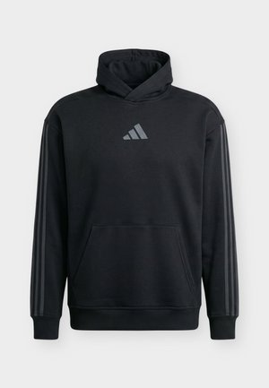 Homme debout face à la caméra, portant un sweat à capuche noir Adidas avec le logo sur la poitrine et un pantalon de jogging gris, sur un fond clair uni.