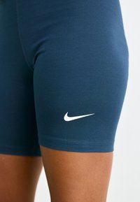Tamsiai mėlynos sportinės šortai, pagaminti iš tamprios medžiagos, su balta Nike logotipo etikete ant šlaunies ir prigludusia forma be siūlių kraštų.