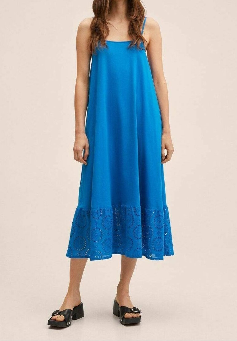 Femme portant une robe midi bleue sans manches avec un ourlet brodé et des sandales compensées noires, se tenant sur un fond neutre.