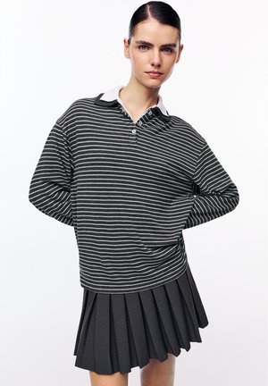 Jonge vrouw met donker, naar achteren gekamd haar, gekleed in een zwart-wit gestreept poloshirt met lange mouwen en een geplooide zwarte rok, tegen een witte achtergrond.