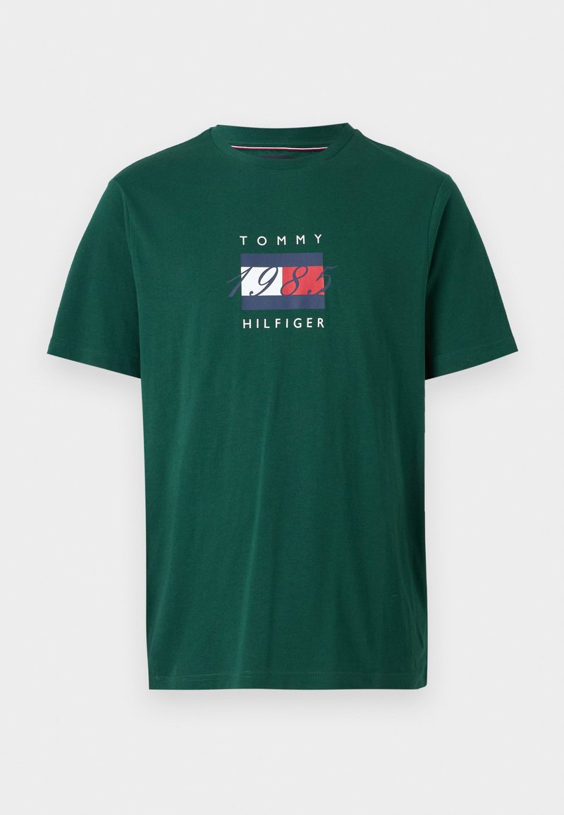 Camiseta de algodón verde con un diseño de logo rectangular que incluye "TOMMY HILFIGER" y "1985" en colores contrastantes. Manga corta y cuello redondo.
