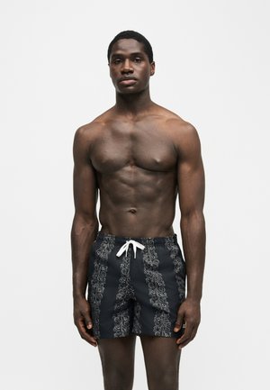 Uomo senza maglietta dalla corporatura muscolosa che indossa pantaloncini da bagno neri con coulisse bianca e motivi a spirale verticali grigio chiaro, in piedi su sfondo bianco.