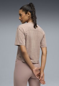 Αθλητικό τοπ με κοντά μανίκια σε ανοιχτό μπεζ χρώμα, με χαλαρή εφαρμογή, στρογγυλεμένο κάτω μέρος και διακριτική λεπτομέρεια με λογότυπο κοντά στο τελείωμα.