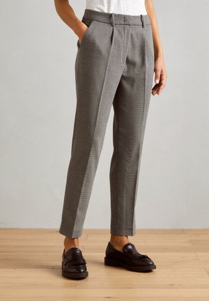 Femme vêtue d’un pantalon gris slim texturé avec poches latérales et de mocassins en cuir noir, debout sur un sol en bois.