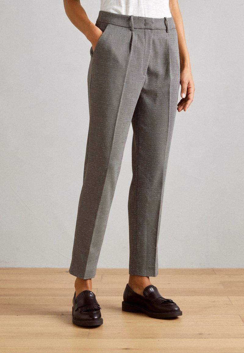 Femme vêtue d’un pantalon gris slim texturé avec poches latérales et de mocassins en cuir noir, debout sur un sol en bois.