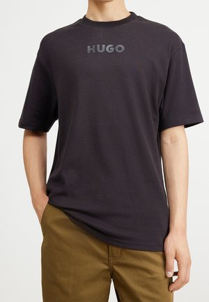 Sort kortærmet t-shirt med struktureret stof og præget "HUGO"-logo på brystet, kombineret med lysebrune bukser.