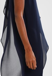 Haut semi-transparent bleu marine avec un design superposé, texture lisse et drapé fluide, doté d'un ourlet incurvé et de côtés ouverts.