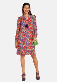 Frau in einem bunten Blumenkleid mit langen Ärmeln, lila Schuhen und grüner Handtasche, die vor einem schlichten weißen Hintergrund lächelt.