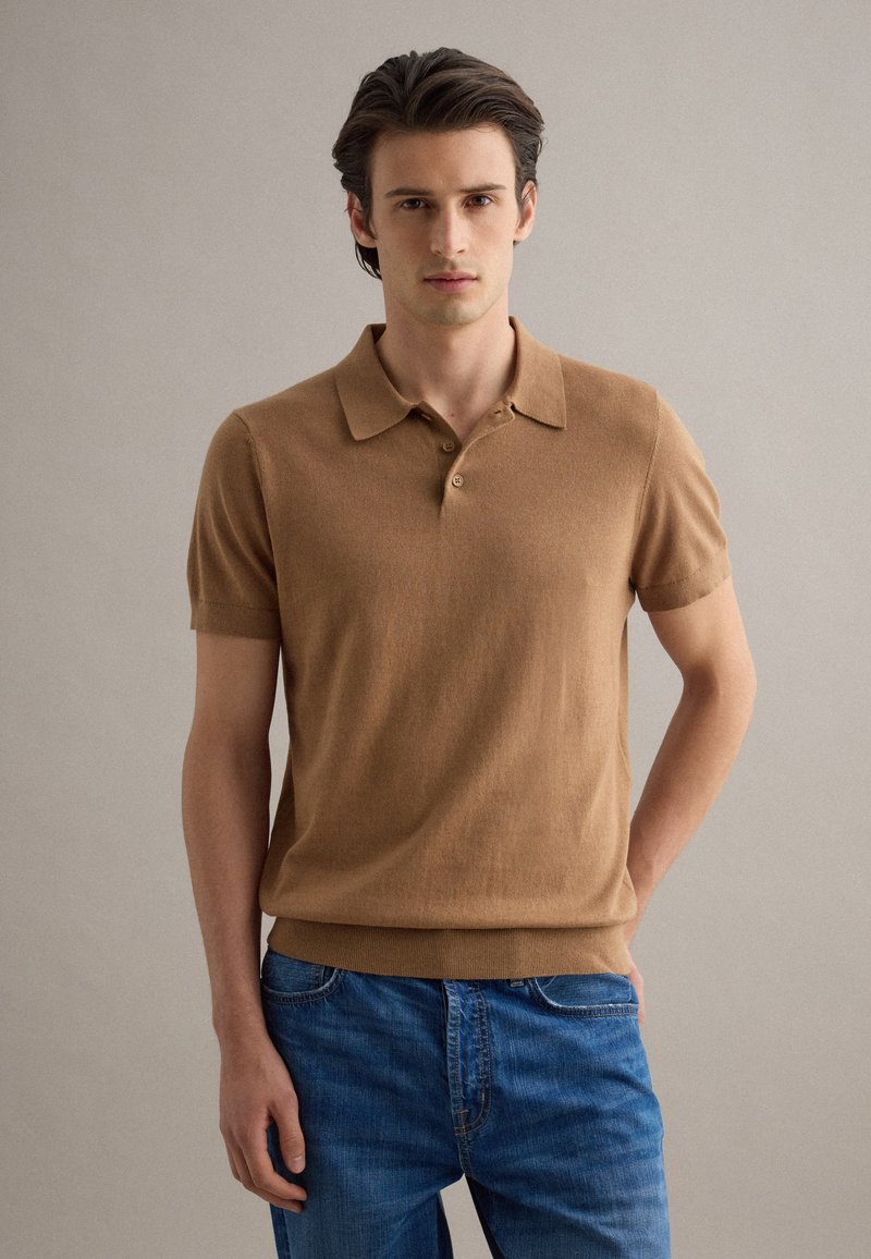 Joven de pie con las manos en los bolsillos, vistiendo una camisa polo de manga corta color camel y pantalones vaqueros azules, frente a un fondo liso.